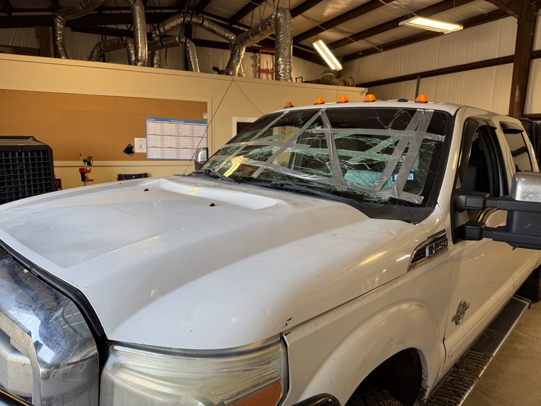 A Ford F-250 with a shattered windshield inside the air-conditioned South Texas Auto Glass shop bay in Corpus Christi, with overhead AC ductwork, where windshield adhesive cures in a clean, cool space